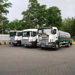 Trzy białe ciężarówki Scania zaparkowane obok siebie na brukowanym parkingu, z drzewami w tle i fragmentem ogrodzenia oraz domu z czerwoną dachówką.