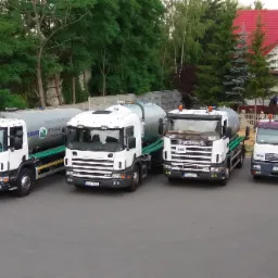 Cztery ciężarówki asenizacyjne różnych marek (Scania i MAN) zaparkowane obok siebie na tle zieleni i domu z czerwoną dachówką. Na cysternach widoczne napisy 'Usługi Asenizacyjne, Wynajem kontenerów...