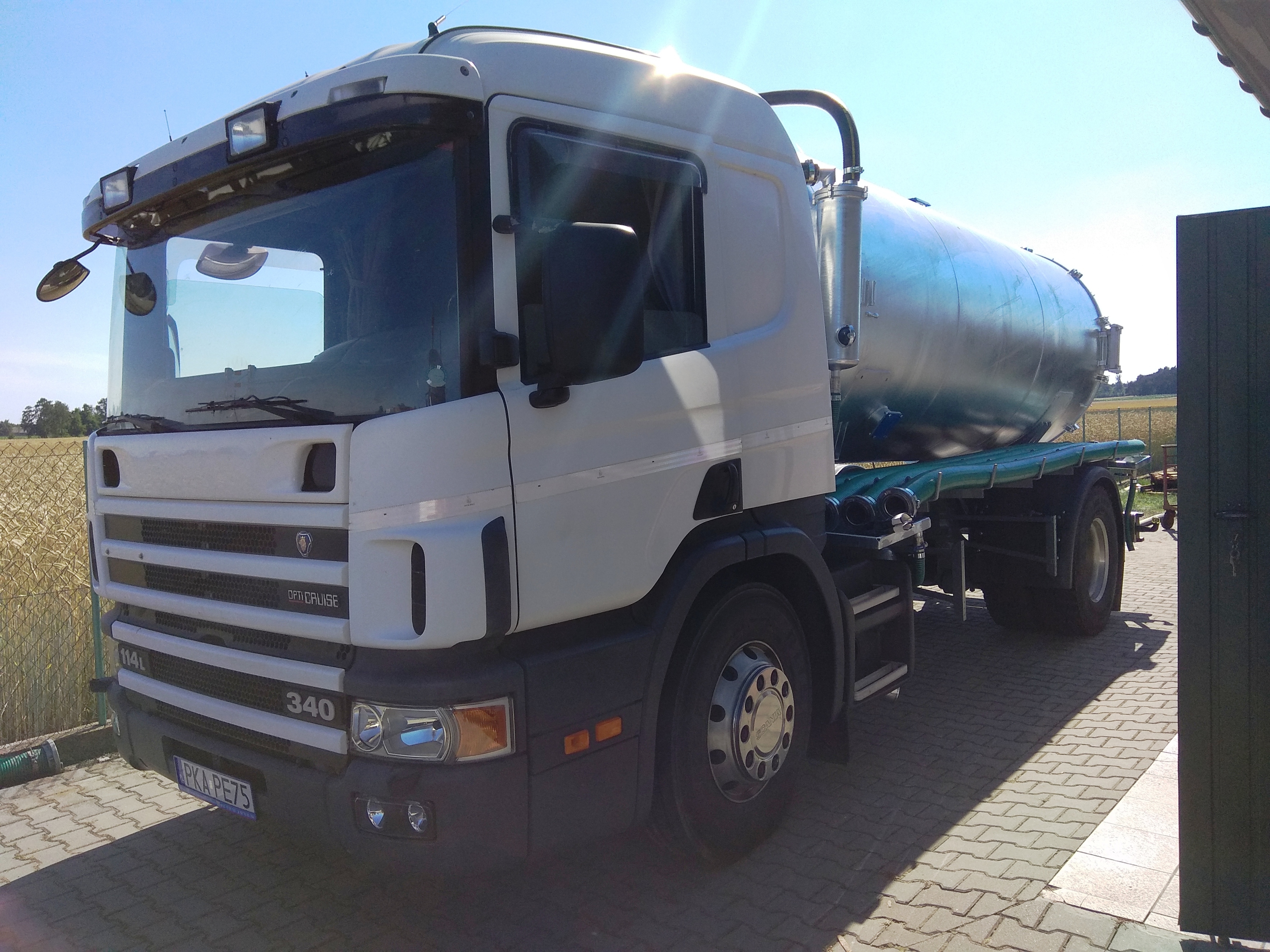 Biała ciężarówka Scania 114L 340 z metalową cysterną na platformie, zaparkowana na kostce brukowej, z polem w tle.