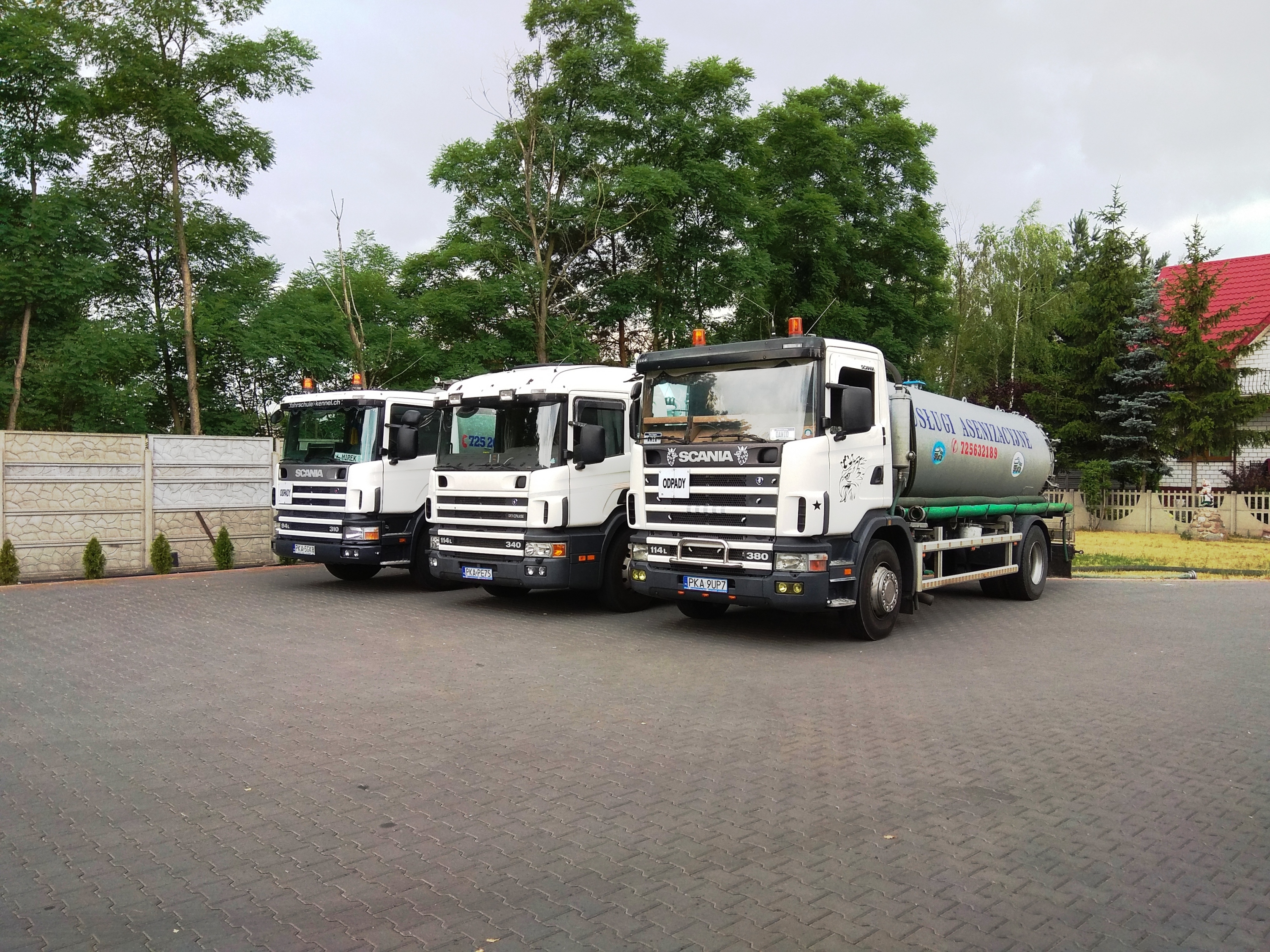Trzy białe ciężarówki Scania zaparkowane obok siebie na brukowanym parkingu, z drzewami w tle i fragmentem ogrodzenia oraz domu z czerwoną dachówką.