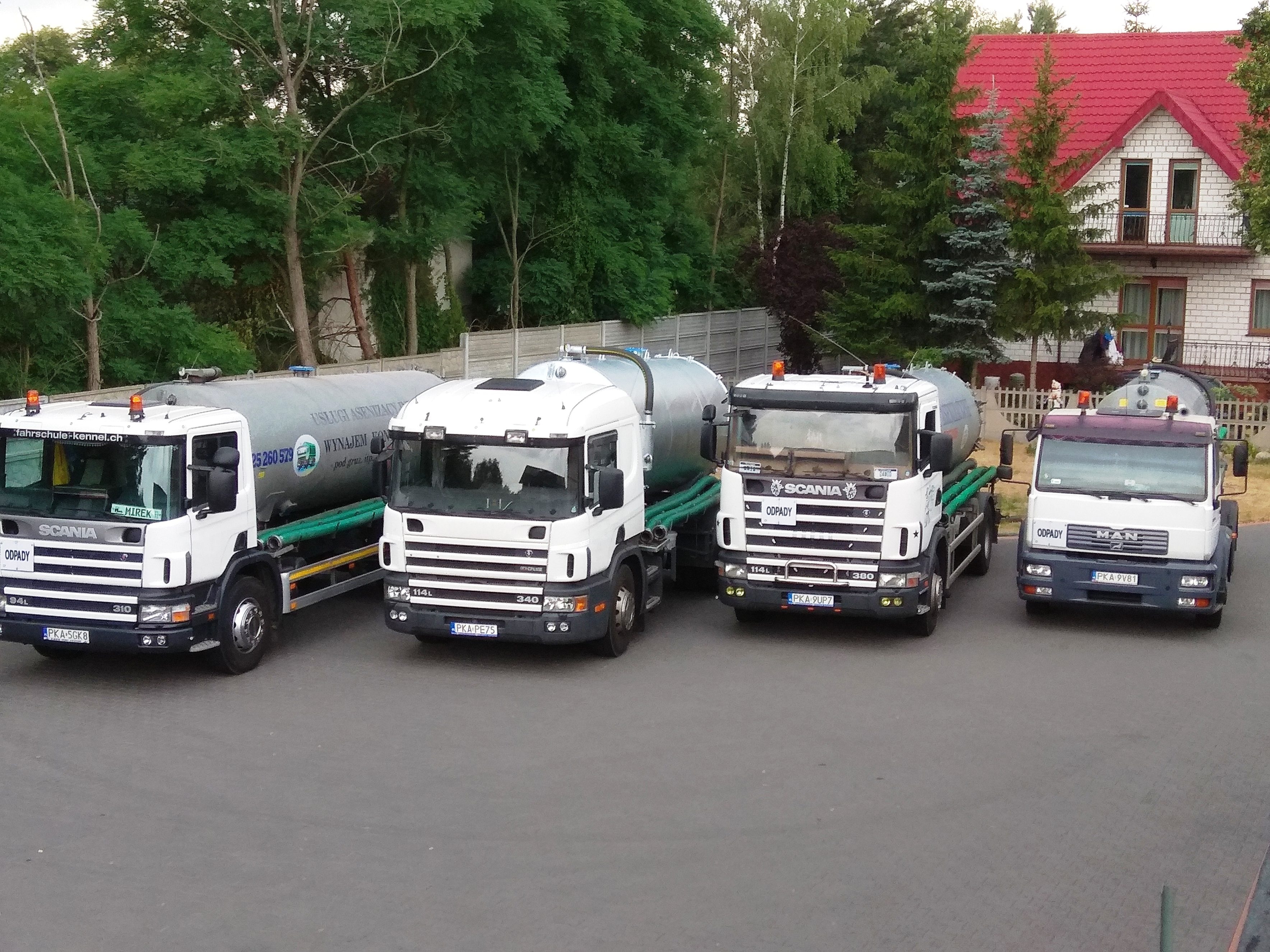 Cztery ciężarówki asenizacyjne różnych marek (Scania i MAN) zaparkowane obok siebie na tle zieleni i domu z czerwoną dachówką. Na cysternach widoczne napisy 'Usługi Asenizacyjne, Wynajem kontenerów...