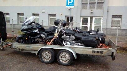 Dwa czarne motocykle zabezpieczone pasami na przyczepie marki Thule, zaparkowanej na tle budynku z szarą elewacją i znakiem parkingowym.