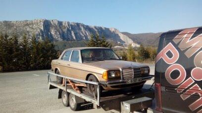 Złoty Mercedes W123 zabezpieczony pasami na lawecie z dwuosiowym podwoziem, z widocznym fragmentem busa z logo firmy transportowej i górskim krajobrazem w tle.