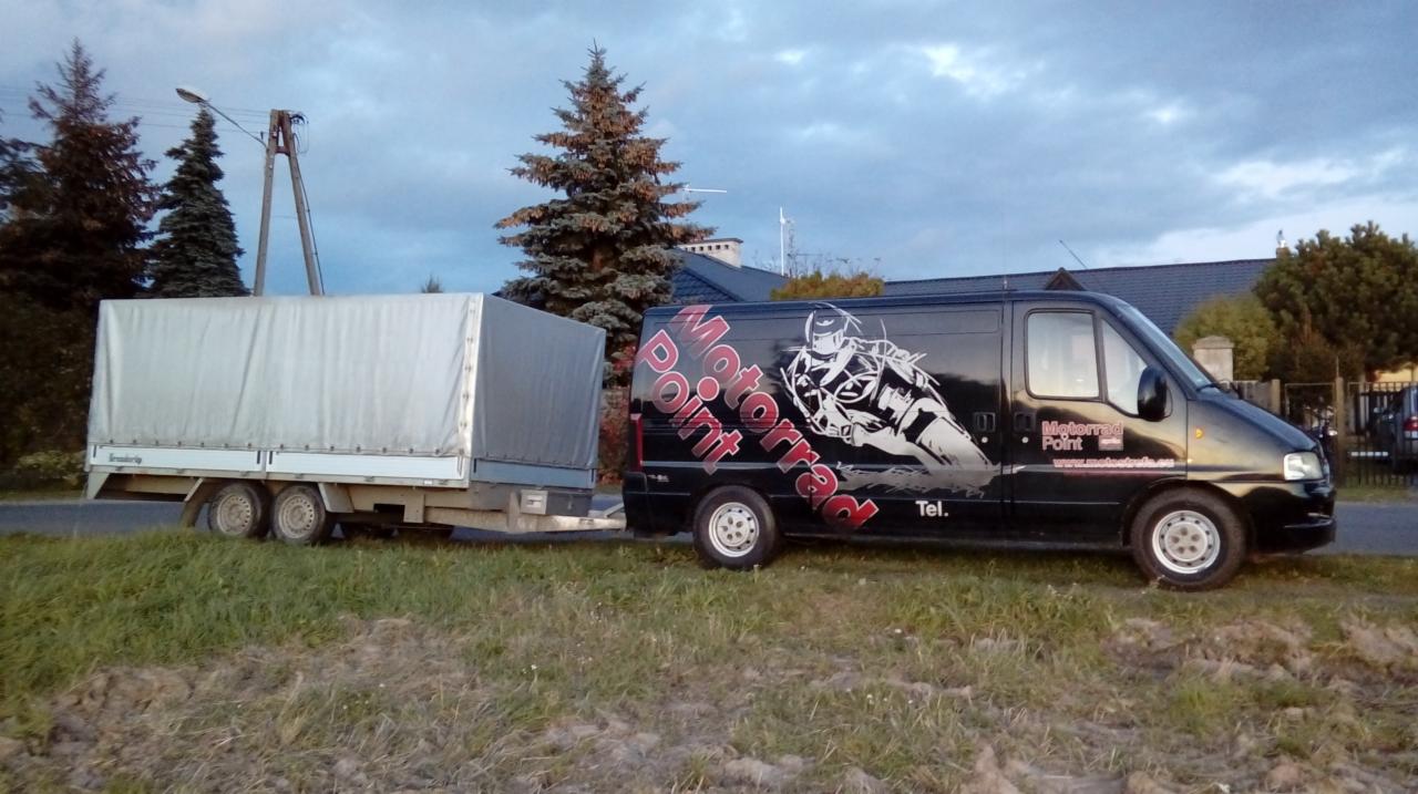 Czarny bus z grafiką motocyklisty i napisem 'Motorrad Point' ciągnie przyczepę z plandeką na tle drzew i pochmurnego nieba.