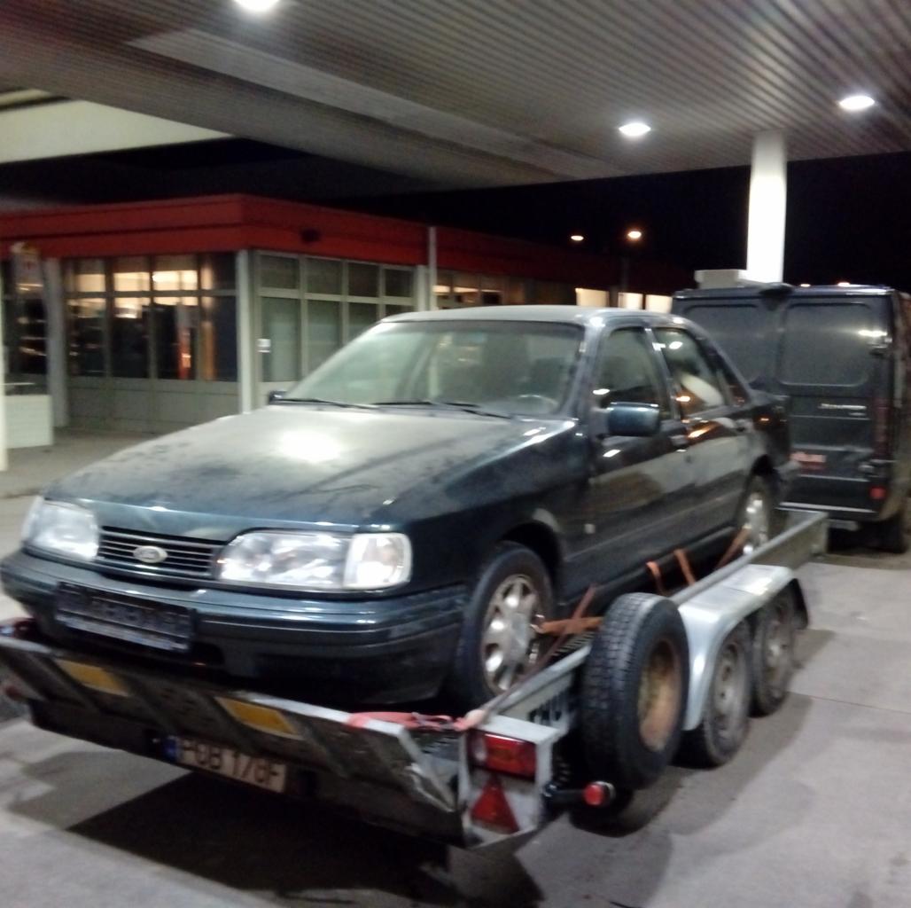 Ciemnozielony sedan Ford Sierra na srebrnej, trzyosiowej lawecie, z czarnym busem w tle, na stacji benzynowej w nocy.