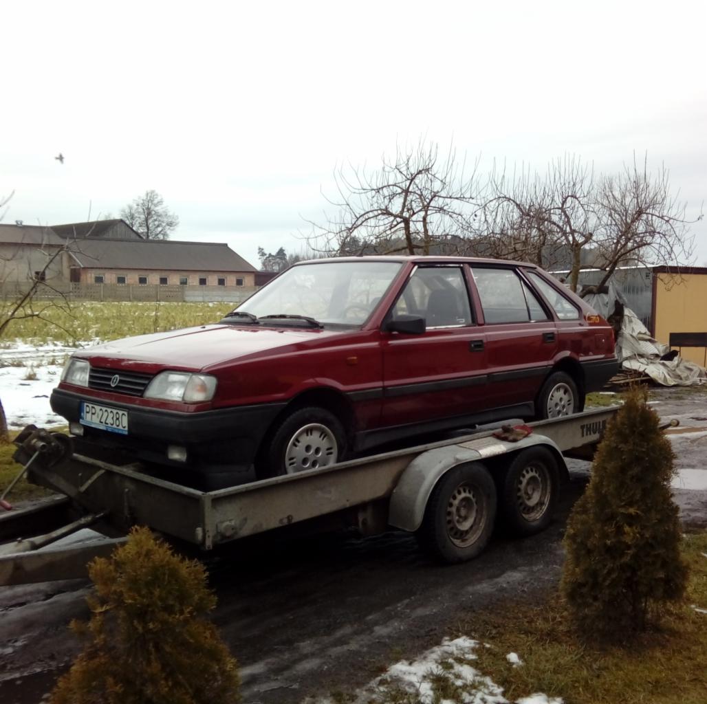 Czerwony samochód FSO Polonez na przyczepie lawety, widoczne tablice rejestracyjne PP 2238C, w tle budynki gospodarcze i drzewa bez liści.