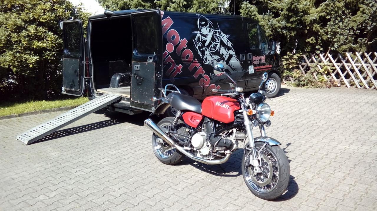 Czerwony motocykl Ducati SuperSport przed czarnym busem z rampą i grafiką motocyklisty na boku.