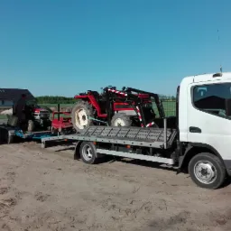 Ciężarówka z platformą przewozi dwa małe, czerwone traktory na tle niebieskiego nieba i domu jednorodzinnego.