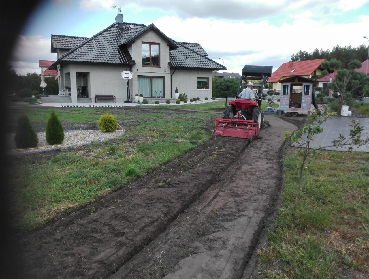 Czerwony traktor z glebogryzarką przygotowuje ziemię pod uprawę na działce z domem jednorodzinnym w tle.