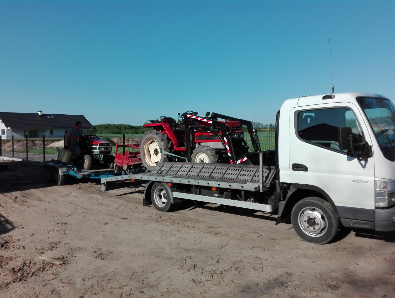 Ciężarówka z platformą przewozi dwa małe, czerwone traktory na tle niebieskiego nieba i domu jednorodzinnego.