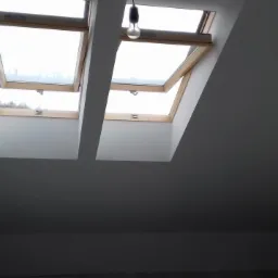 Dwa okna dachowe w pomieszczeniu po remoncie w Dziergowicach, ściany pomalowane na szaro, widoczne niedoskonałości tynku i wisząca żarówka.