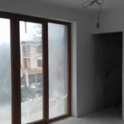 Surowe wnętrze w trakcie wykańczania, z dużym oknem balkonowym z drewnianą ramą, widok na budynek z balkonami i zieleń na zewnątrz, ściany świeżo pokryte gładzią, widoczne punkty elektryczne...