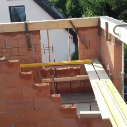 Budowa ścian z czerwonej cegły, widoczne drewniane szalunki i poziomica, dom w tle.