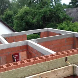 Surowy stan budynku z czerwonej cegły i betonu, widoczne zbrojenia i izolacja na dachu, w tle domy jednorodzinne i drzewa.