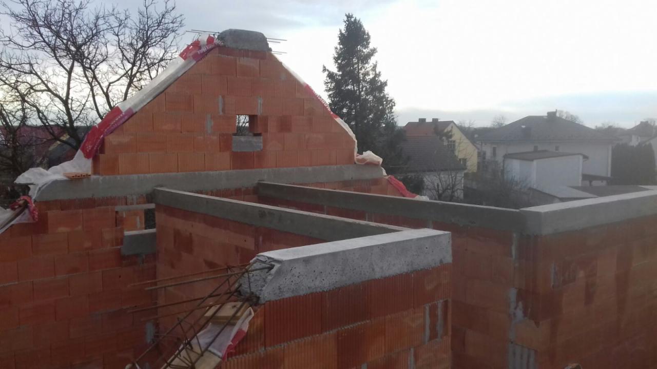 Wznoszenie murów z czerwonej cegły ceramicznej z widocznymi elementami żelbetowymi, zbrojeniami i zabezpieczeniem folią na szczycie, w tle zabudowa jednorodzinna.