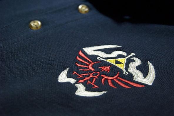 Granatowa koszulka polo z haftowanym motywem inspirowanym grą The Legend of Zelda, przedstawiającym czerwoną hyruleńską tarczę z białymi i żółtymi akcentami, widoczne dwa złote guziki.