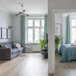 Jasne mieszkanie z drewnianą podłogą, szarą sofą z pastelowymi poduszkami, białym stołem i widokiem na sypialnię z łóżkiem w błękitnej narzucie przez otwarte drzwi.