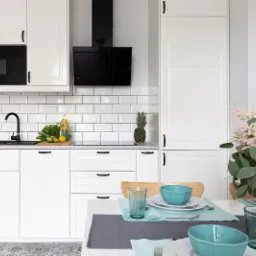 Jasna kuchnia z białymi szafkami, czarną armaturą i aneksem jadalnym z turkusową zastawą i różowym wazonem z kwiatami.
