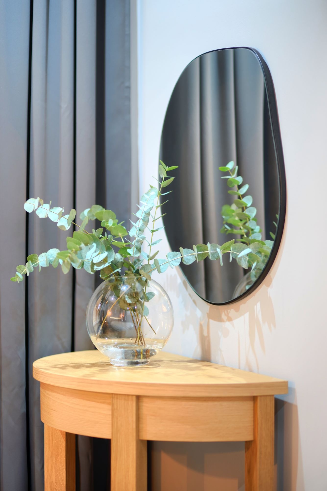 Okrągłe lustro z czarną ramą zawieszone nad półokrągłym, jasnym, drewnianym stolikiem, na którym stoi szklany wazon z eukaliptusem, obok zasłona w odcieniach szarości.