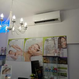 Cool-Install - Biały klimatyzator ścienny zamontowany w salonie kosmetycznym nad cennikami usług, oświetlony przez ozdobny, biały żyrandol.