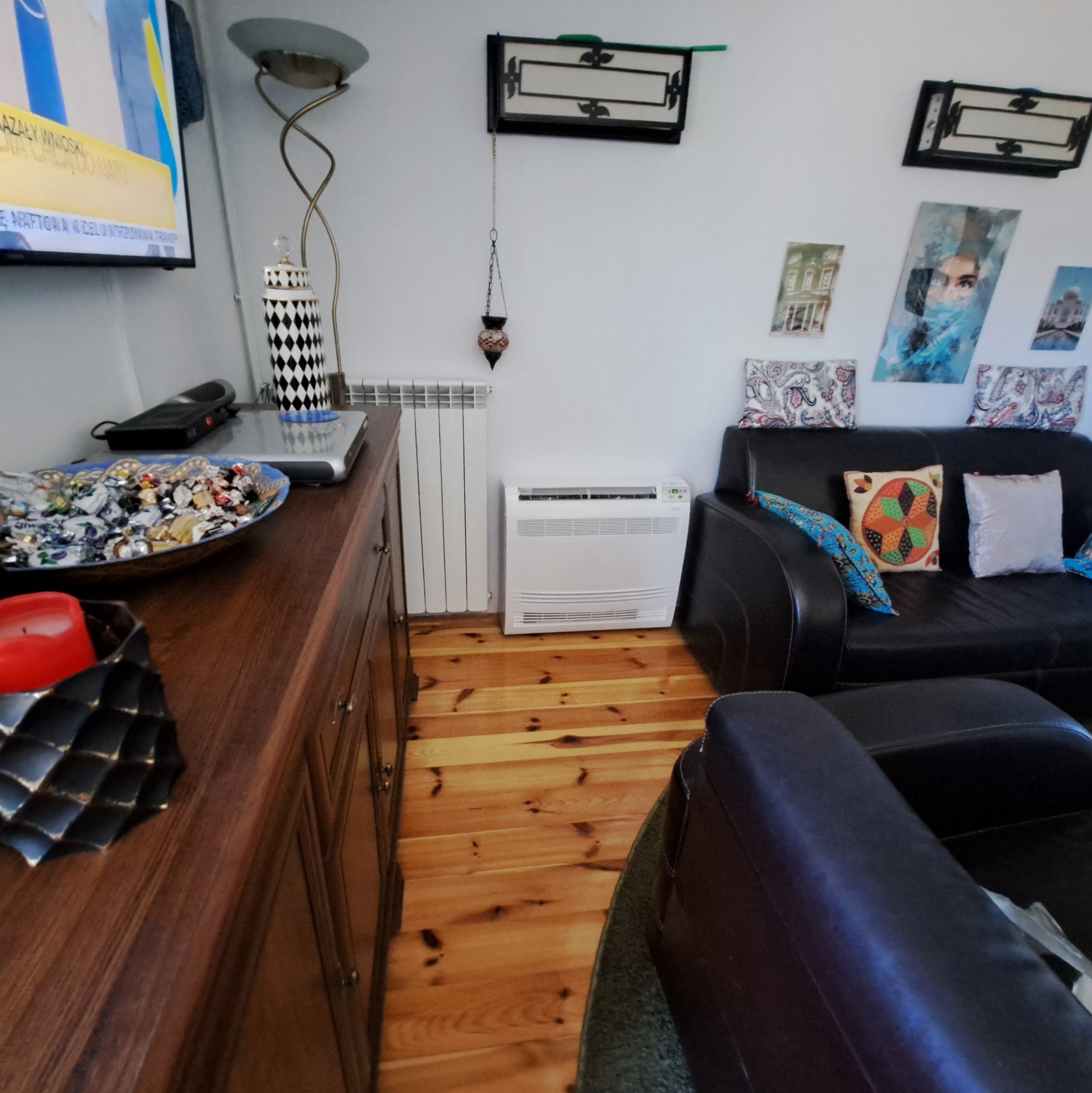 Biała jednostka konsolowa klimatyzatora Rotenso ustawiona na drewnianej podłodze przy grzejniku i ciemnej szafce z dekoracyjną misą słodyczy, w tle czarna sofa z poduszkami i ozdobne obrazy...