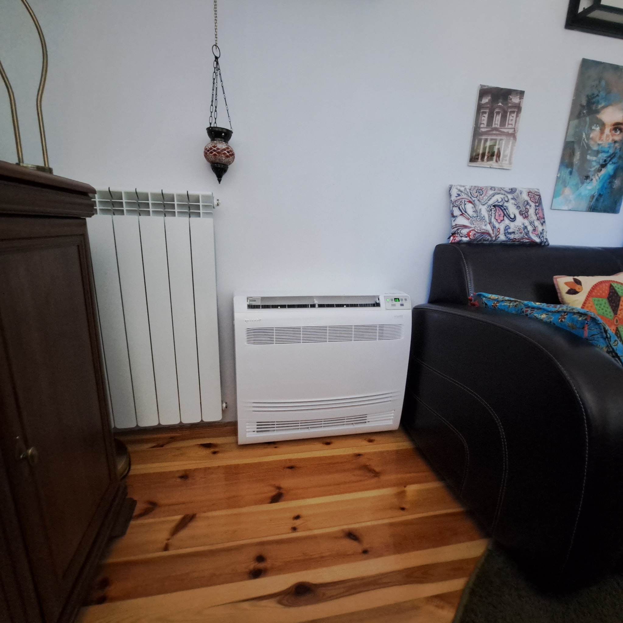 Wolnostojący klimatyzator pokojowy Rotenso Imoto+ ustawiony na drewnianej podłodze w salonie, obok grzejnika i skórzanej sofy, z widoczną dekoracyjną lampą wiszącą.