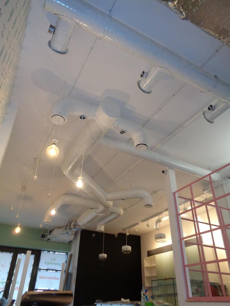 Białe kanały wentylacyjne zamontowane pod sufitem w lokalu w Warszawie, widoczne również lampy i fragmenty wyposażenia.