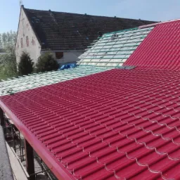 Czerwona blachodachówka na nowym dachu w Paczkowie, w tle budynek z ciemnym, starszym pokryciem i przygotowane łaty pod kolejny fragment poszycia.