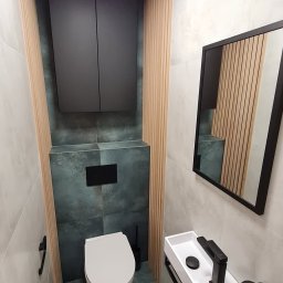 STYLOWE WNĘTRZE WARSZAWA MARIUSZ BIERNACKI - Nowoczesna łazienka z wiszącą toaletą, umywalką nablatową i drewnianymi lamelami na ścianach. Ciemna szafka nad toaletą i lustro w czarnej ramie.