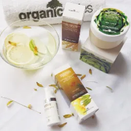 Kompozycja produktów organicznych do pielęgnacji ciała: nawilżający krem, mus do ciała, tonik rozjaśniający oraz miska z wodą i plasterkami cytryny i limonki na białym ręczniku z logo.