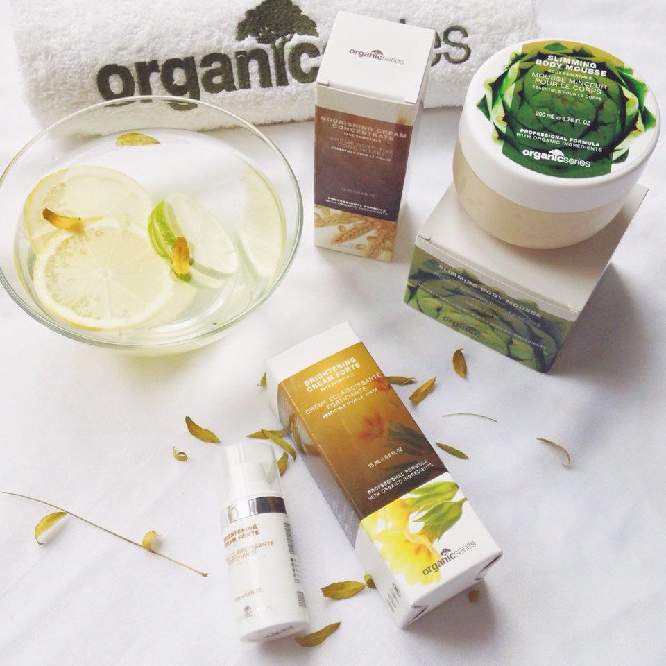 Kompozycja produktów organicznych do pielęgnacji ciała: nawilżający krem, mus do ciała, tonik rozjaśniający oraz miska z wodą i plasterkami cytryny i limonki na białym ręczniku z logo.