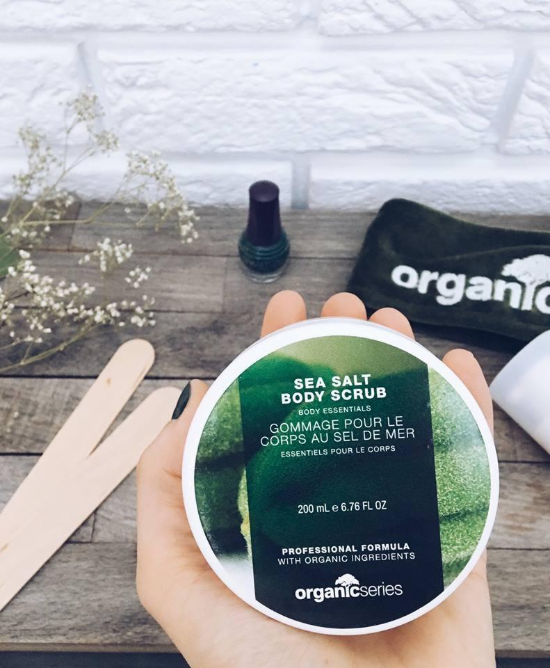 Dłoń trzyma okrągłe opakowanie na peeling solny do ciała Organic Series na tle drewnianego blatu z dekoracjami: lakier do paznokci w odcieniu butelkowej zieleni, opaska na włosy z logo organic...