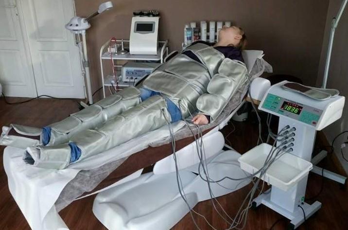 Kobieta leżąca na łóżku zabiegowym, ubrana w kombinezon do presoterapii, widoczny panel sterowania urządzenia z wyświetlaną wartością 1836, w tle sprzęt kosmetyczny.