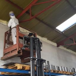 Termoprof Dawid Osiński - Pracownik w kombinezonie ochronnym aplikuje izolację natryskową na ścianę hali przemysłowej z podnośnika koszowego; widoczna metalowa konstrukcja dachu z oknami dachowymi i regały magazynowe.