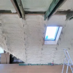 Poddasze po natrysku izolacji pianką, widoczne krokwie, okno dachowe i drabina, szara rura wentylacyjna przy podłodze.