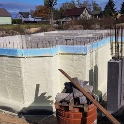 Termoprof Dawid Osiński - Fundament budynku w trakcie izolacji: robotnik w kombinezonie ochronnym aplikuje warstwę izolacyjną na ścianę fundamentową, widoczne zbrojenia i niebieskie płyty izolacyjne, w tle zabudowa...