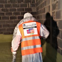 Termoprof Dawid Osiński - Pracownik w kombinezonie ochronnym i pomarańczowej kamizelce z logo firmy izolacyjnej, trzymający w rękach owiniętą rurę, stoi w narożniku pomieszczenia z ciemnymi, surowymi ścianami z cegły.