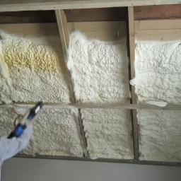 Termoprof Dawid Osiński - Pracownik w kombinezonie ochronnym aplikuje pianę izolacyjną na poddaszu, widoczne drewniane krokwie i częściowo zaizolowana powierzchnia z żółtymi przebarwieniami na piance.