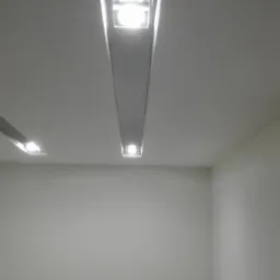 Zamontowane podłużne lampy LED w suficie podwieszanym z płyt karton-gips, oświetlające białe ściany w pomieszczeniu.