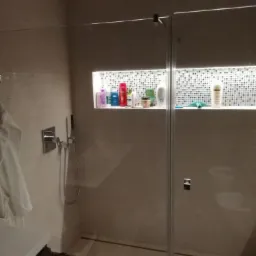 Nowoczesna kabina prysznicowa z wnęką wyłożoną mozaiką, podświetloną LED i wypełnioną kosmetykami, widoczny chromowany zestaw prysznicowy i fragment umywalki z ręcznikiem.