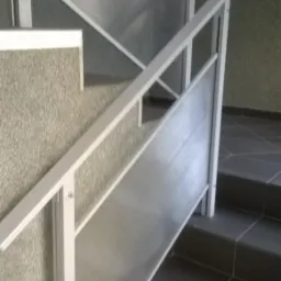 Wewnętrzna klatka schodowa z szarą, teksturowaną ścianą i nowoczesną, minimalistyczną balustradą z białych profili i wypełnieniem z półprzezroczystego tworzywa sztucznego w pionowe prążki.