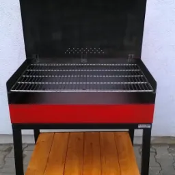 Otwarty, metalowy grill z czerwonym akcentem i drewnianą półką na kółkach, stojący na brukowanym podłożu.