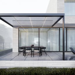 NUVIO Architects sp. z o.o - Nowoczesny taras z pergolą i minimalistycznym zestawem mebli ogrodowych na tle białej, ceglanej elewacji domu w mglisty dzień.