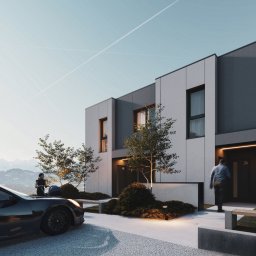 NUVIO Architects sp. z o.o - Nowoczesne segmenty mieszkalne z szarą elewacją, oświetlonym wejściem i zagospodarowanym terenem zielonym. W tle góry i osoba z wózkiem przy samochodzie.