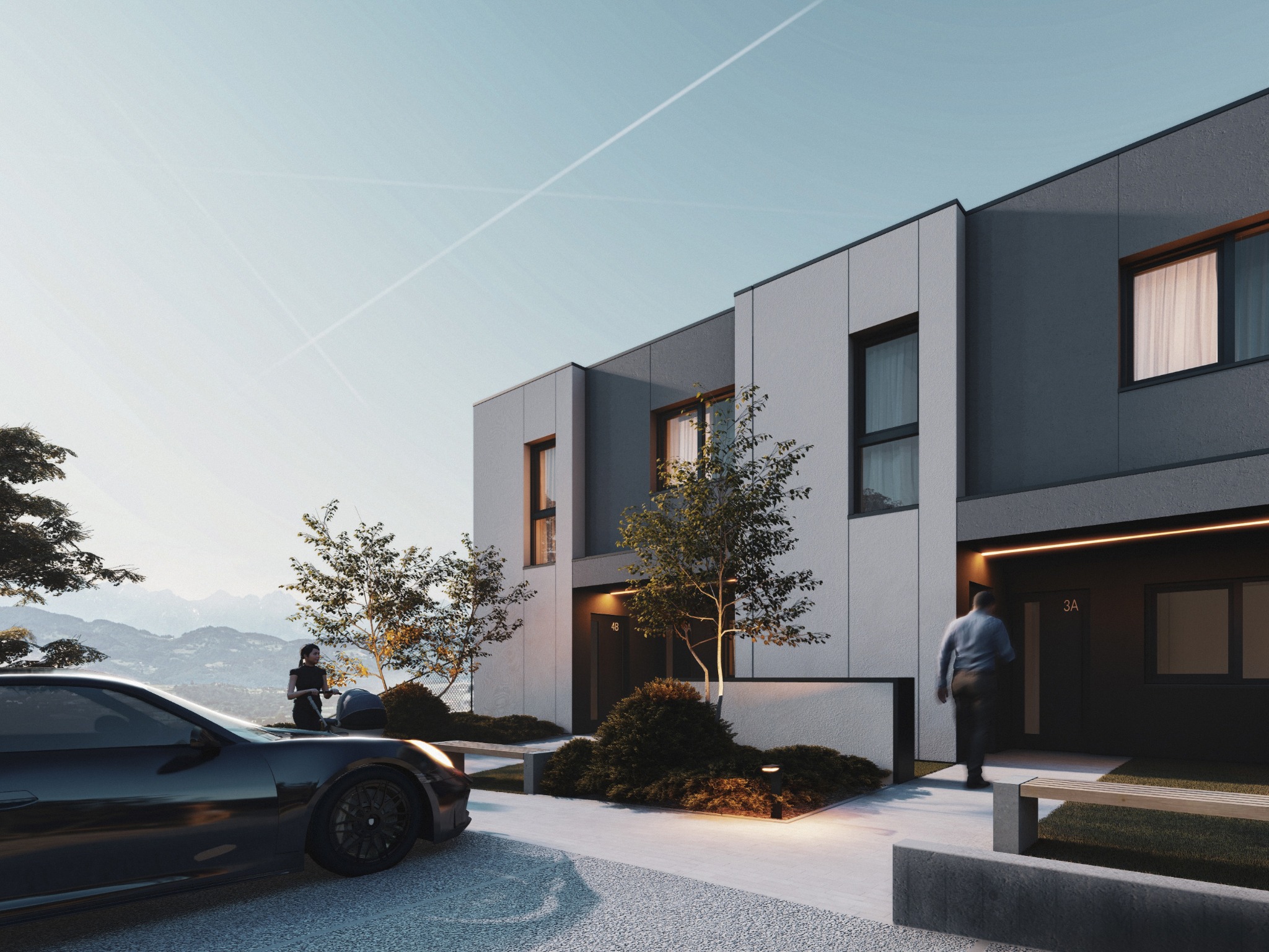 Nowoczesne segmenty mieszkalne z szarą elewacją, oświetlonym wejściem i zagospodarowanym terenem zielonym. W tle góry i osoba z wózkiem przy samochodzie.