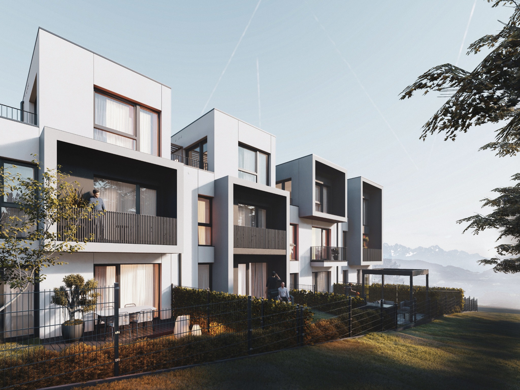 Nowoczesne, białe budynki wielorodzinne z balkonami i tarasami, otoczone zielenią i ogrodzeniem. W tle widoczne góry i błękitne niebo z smugami kondensacyjnymi.