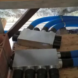 Dwa metalowe rozdzielacze z podłączonymi niebieskimi, karbowanymi przewodami wentylacyjnymi, zamontowane na drewnianej konstrukcji poddasza z widoczną izolacją piankową.