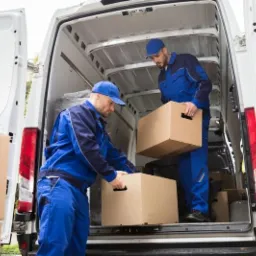 Dwóch mężczyzn w niebieskich kombinezonach umieszcza kartonowe pudła wewnątrz białego furgonetki z otwartymi drzwiami bagażnika, widoczne światła stopu, pudła z uchwytami na dłonie.