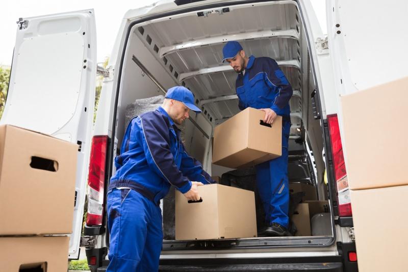 Dwóch mężczyzn w niebieskich kombinezonach umieszcza kartonowe pudła wewnątrz białego furgonetki z otwartymi drzwiami bagażnika, widoczne światła stopu, pudła z uchwytami na dłonie.