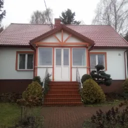 Dom jednorodzinny z białymi ścianami, czerwoną dachówką i pomarańczowymi elementami wykończenia, widok z przodu na wejście z ceglanymi schodami i metalowymi poręczami, otoczony zielenią.